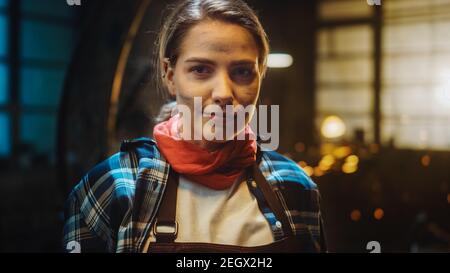 Giovane bella Empowerering Donna con l'orecchio Piercing sorride delicatamente alla macchina fotografica. Autentico Fabricator che indossa abiti da lavoro in un laboratorio di metallo. Foto Stock