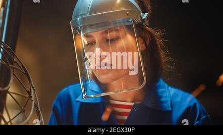 Primo piano Ritratto di bella Femminile Fabricator in Jumpsuit blu e maschera di sicurezza. Sta macinando una scultura in metallo con una smerigliatrice angolare in uno studio Foto Stock