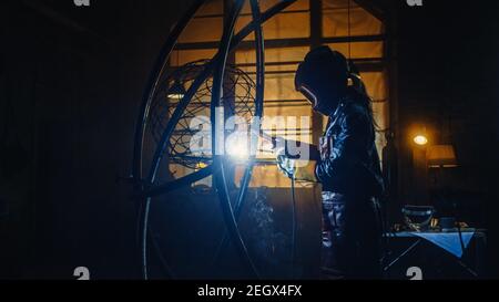 Giovane artista femminile contemporanea sta saldando la sua scultura di tubo metallico con un attrezzo di potenza portatile in un laboratorio di studio. Emancipazione Donna rende moderno Foto Stock