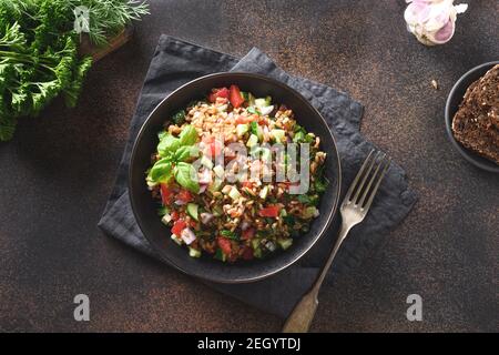 Farro di cereali interi cotti come insalata con verdure, pomodori, cetrioli, verdi su fondo marrone. Sana cena vegana. Vista dall'alto. Foto Stock