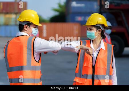 Assistente asiatico nuovo saluto normale da gomiti urti indossare sicurezza casco e maschera nel terminale del deposito del contenitore per prevenire il coronavirus e la distanza sociale Foto Stock