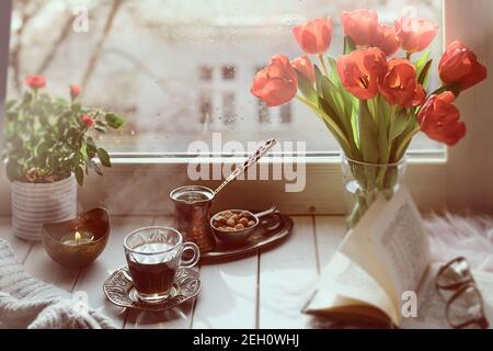 Caffè orientale in tradizionale caffettiera turca di rame con fiori sul davanzale della finestra. Rustico davanzale in legno con mazzo di libro tulipani. Pioggia fredda Foto Stock