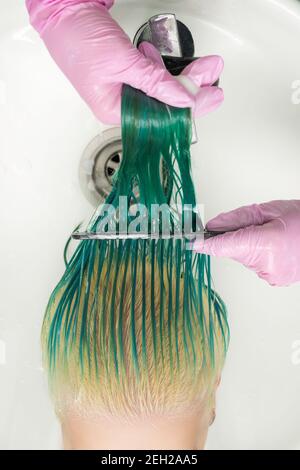 Foto ad alto angolo del parrucchiere in un guanto protettivo che tiene i capelli bagnati nelle mani e pettina i capelli verdi lunghi e scoloriti della donna, mentre shampooing i capelli Foto Stock