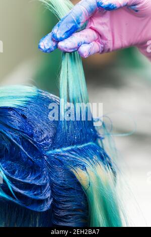 Parrucchieri con mano in guanto protettivo solleva shock di moderni capelli blu del cliente durante il processo di tintura dei capelli in salone di bellezza professionale. Foto Stock