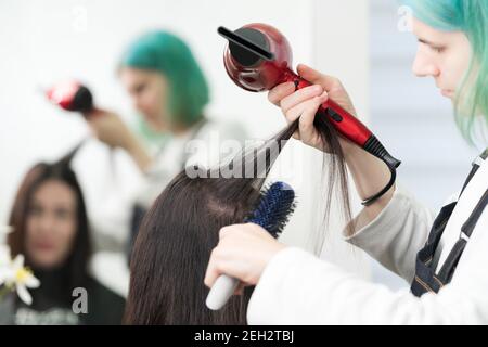 Il parrucchiere femminile asciuga i capelli della brunetta con asciugacapelli rosso e spazzola blu nel salone di bellezza professionale. Foto Stock