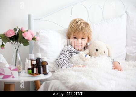 Malato piccolo biondo bambino, sdraiato a letto con febbre, abbracciando piccolo peloso giocattolo, medicina accanto a lui sul comodino Foto Stock