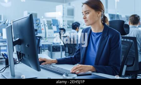 Modern Factory Office: Ritratto di giovane e fiduciosa ingegnere industriale femminile che lavora su computer Holding Metal Component e ispettendolo. Grande Foto Stock