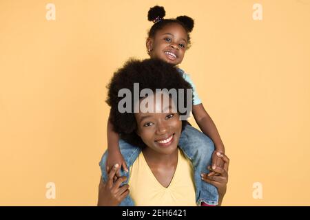 Ritratto di felice bambina africana seduta sulle spalle della mamma e divertendosi, ridendo alla macchina fotografica su sfondo giallo isolato Foto Stock
