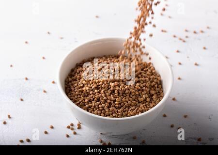 I grani di grano saraceno cadono in una ciotola di ceramica bianca su uno sfondo grigio chiaro. Cibo sano e naturale Foto Stock