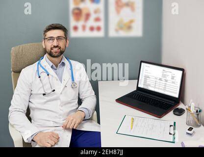 Medico esperto seduto sul posto di lavoro con un cappotto medico durante l'analisi della cartella clinica del paziente Foto Stock