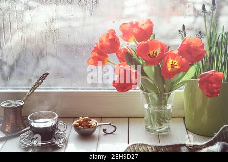 Caffè orientale in tradizionale caffettiera turca di rame con fiori sul davanzale della finestra. Davanzale in legno con mazzo di libro tulipani. Giorno freddo piovoso in s. Foto Stock