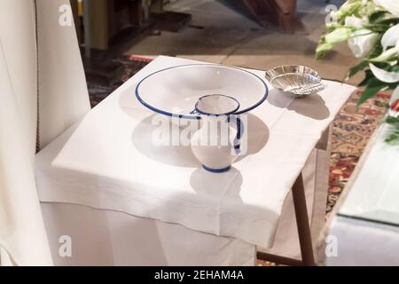 Oggetti di battesimo preparati su un tavolo all'interno di una chiesa. Caraffa con acqua benedetta, lavandino e guscio. Foto Stock