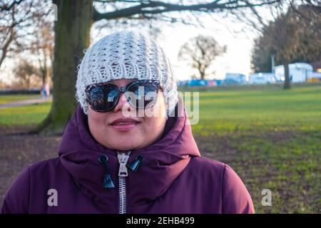 Ritratto di una signora asiatica che indossa un caldo cappotto invernale, cappello e occhiali da sole in un parco alla luce del sole nel tardo pomeriggio in un giorno nel febbraio 2021. Foto Stock