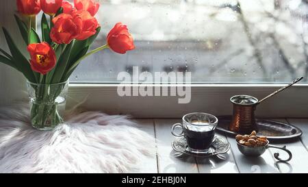 Caffè orientale in tradizionale caffettiera turca di rame con fiori sul davanzale della finestra. Rustico davanzale in legno con mazzo di tulipani e libro. Freddo Foto Stock