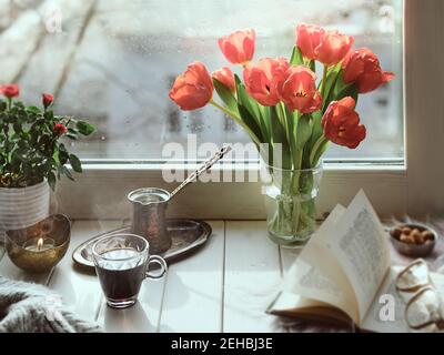 Caffè orientale in tradizionale caffettiera turca di rame con fiori sul davanzale della finestra. Rustico davanzale in legno con mazzo di tulipani e libro. Freddo Foto Stock