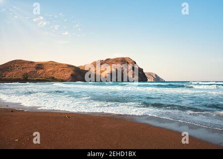 Spiaggia di Las Negras al tramonto, vista sull'oceano e alcune onde. Montagne del deserto sullo sfondo. Cielo blu con alcuni soffici raggi caldi del sole. Foto Stock