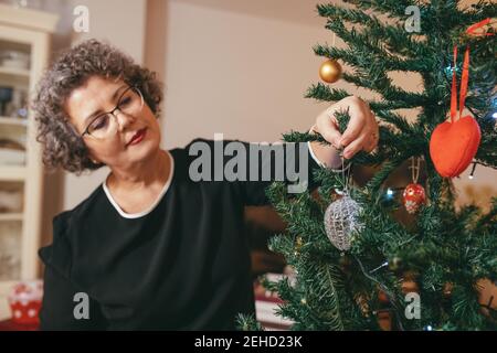Donna di media età focalizzata negli occhiali con decorazione a testa inclinata Abete prima di Capodanno in casa Foto Stock