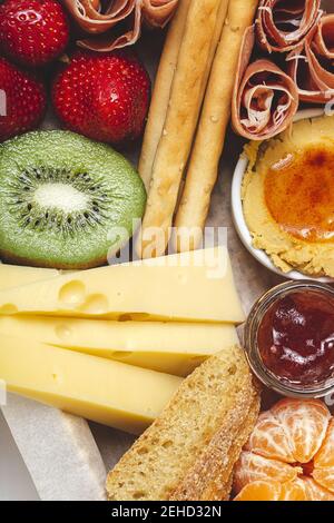 Da sopra scatola del brunch closeup con varie carni a fette assortite tipi di formaggio e croccanti disposti vicino kiwi tazza matura fragole dolci e pe Foto Stock