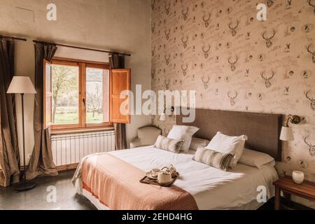 Interno di elegante camera da letto con comodo letto coperto di copriletto vicino alla finestra decorata con tende Foto Stock
