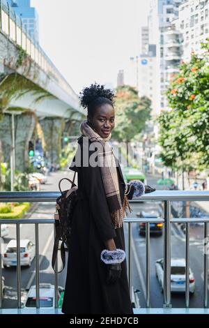 Vista laterale di una giovane femmina nera sorridente in un abbigliamento elegante con zaino che guarda la fotocamera dal ponte sopra la strada urbana Foto Stock
