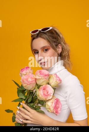 Giovane bella donna in vestito bianco e occhiali da sole alla moda tenuta rosa delicato mentre si sta su sfondo giallo nella foto studio Foto Stock