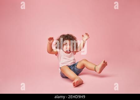 Cute ragazza allegra del toddler con i capelli ricci in vestiti casual divertirsi a fare i volti mentre si siede sul pavimento guardando via su sfondo rosa Foto Stock