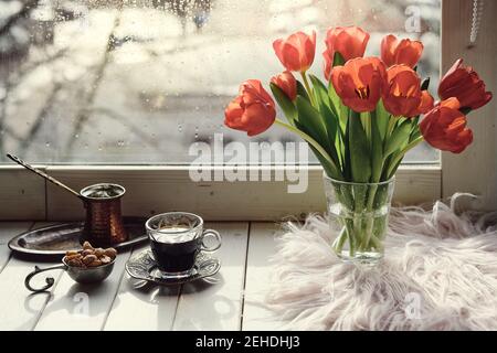 Caffè orientale in tradizionale caffettiera turca di rame con fiori sul davanzale della finestra. Rustico davanzale in legno con mazzo di tulipani e libro. Freddo Foto Stock