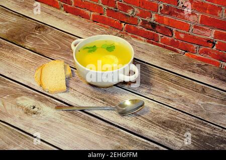 Piatto rotondo profondo con manici di brodo di pollo fresco con uovo, cucchiaio e due fette di pane bianco su un tavolo di legno contro un muro di mattoni rossi. Chiudi-u Foto Stock