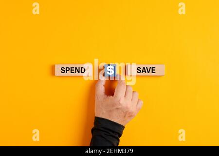 La mano maschile tiene un cubo di legno con il simbolo del dollaro USA tra le opzioni di spesa o di salvataggio su sfondo giallo. Possibilità di scegliere tra spendere o risparmiare Foto Stock