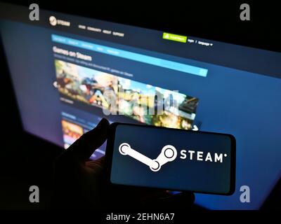 Persona in possesso di smartphone con logo della piattaforma di distribuzione Steam gestito da Valve sullo schermo davanti al sito Web. Mettere a fuoco il display del telefono. Foto Stock