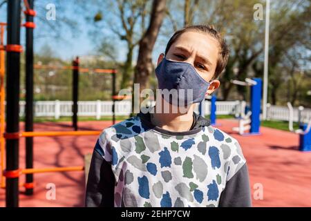 Un ragazzo adolescente in una maschera medica, in piedi sulla strada in un parco e schiaccato, barre orizzontali sullo sfondo. Giorno di sole. Concetto di coronavirus, Foto Stock