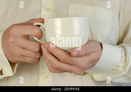 L'uomo adulto sta tenendo una grande tazza bianca. Un uomo caucasico di mezza età in una camicia crema con gemelli. All'interno. Messa a fuoco selettiva. Foto Stock
