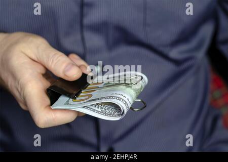 Un uomo in una camicia blu tiene fuori i dollari serrati con una clip del raccoglitore. Diverse centinaia di banconote da dollaro si sono piegate a metà in una mano dell'uomo. Uomo di mezza età, Foto Stock