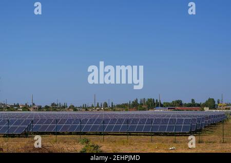 File di pannelli solari dietro una recinzione in un campo. Una centrale solare contro lo sfondo di una piccola comunità residenziale. Messa a fuoco selettiva. Foto Stock