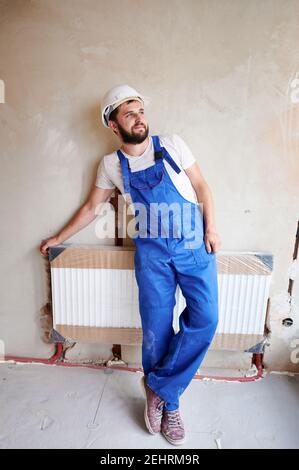 Giovane idraulico bearded, indossando tute blu, casco bianco, in piedi, poggiato su appena installato radiatore d'acqua, sorridente, soddisfatto. Verticale. Concetto di costruzione e ristrutturazione domestica. Foto Stock