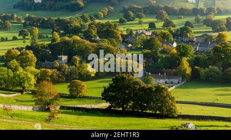 Sunlit pittoresco villaggio Dales (chiesa e case) annidato in valle da collina e alberi in colori autunnali - Arncliffe, Yorkshire Dales, Inghilterra, Regno Unito Foto Stock