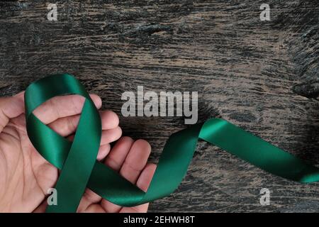 Vista dall'alto del nastro verde smeraldo che tiene la mano su sfondo di legno scuro. Concetto di consapevolezza del cancro del fegato. Foto Stock