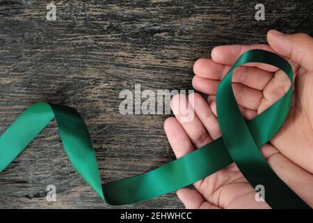 Vista dall'alto del nastro verde smeraldo che tiene la mano su sfondo di legno scuro. Concetto di consapevolezza del cancro del fegato. Foto Stock