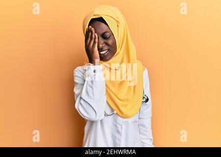 Bella giovane donna africana che indossa uniforme medico e hijab sbadiglio stanco coprendo metà faccia, occhio e bocca con la mano. Faccia fa male nel dolore. Foto Stock