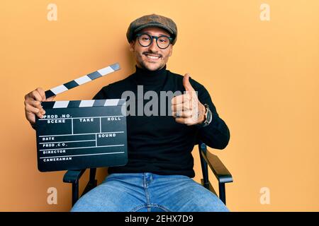 Bell'uomo con tatuaggi che tengono clapboard video film seduto sulla sedia regista sorridente felice e positivo, pollice su facendo eccellente e sigg approvazione Foto Stock