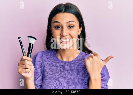 Giovane donna ispanica che tiene le spazzole di trucco che punta il pollice fino a. il lato sorridente felice con bocca aperta Foto Stock