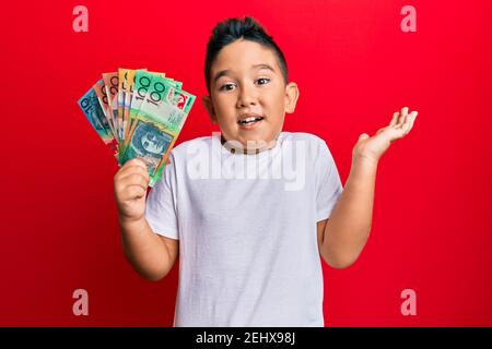 Piccolo ragazzo ispanico capretto che tiene i dollari australiani che festeggia il successo con buon sorriso e espressione vincente con mano sollevata Foto Stock