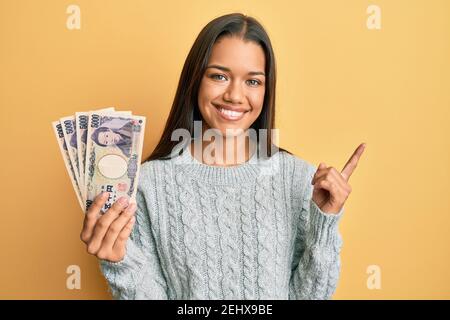 Bella donna ispanica che tiene 5000 yen giapponesi banconote sorridente felice puntare con la mano e il dito verso il lato Foto Stock