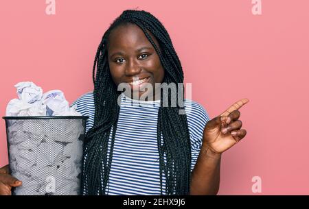 Giovane donna nera con trecce che tengono il bidone di carta pieno di carta sminuzzata sorridente che punta felice con la mano e il dito a. il lato Foto Stock