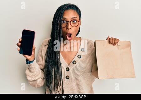 Donna afroamericana che tiene il sacchetto di carta di consegna e che mostra lo schermo dello smartphone impaurito e scioccato con sorpresa e stupito espressione, paura e scossa Foto Stock