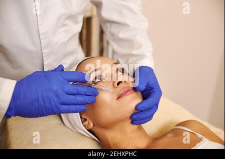 Closeup di una donna cute viso e mani estetiste in guanti medici blu di protezione che tengono siringa con prodotto di bellezza vicino al viso. Sollevamento del viso Foto Stock