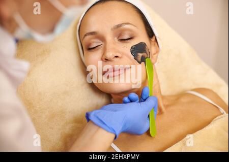 Closeup del viso di bella donna e mani esteticista che applicano un prodotto di bellezza nero sulla sua faccia Foto Stock