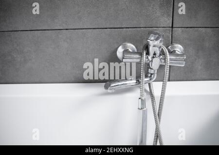 In un bagno con piastrelle grigie, troverete un rubinetto cromato con doccia e vasca bianca. Elegante bagno bianco con rubinetto d'argento. Minimalismo all'interno del bagno. Foto Stock