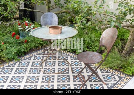 Mobili da giardino con tavolo vintage in metallo e sedie su patio piastrellato mosaico pavimentato con alberi di mele dietro un muro reso Inghilterra GB UK Foto Stock