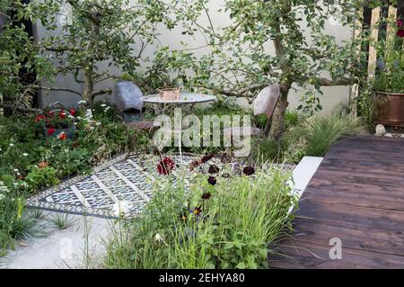 Cortile giardino tavolo e sedie su patio mosaico piastrellato con alberi da frutta di mela contro una parete giardino renderizzata terrazza in legno in estate Inghilterra UK Foto Stock